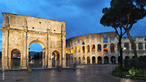 The iconic Colosseum and the Arch of Constantine beautifully illuminated against the dramatic blue hour sky in Rome, Italy. An ancient and majestic historical landmark, a popular tourist destination