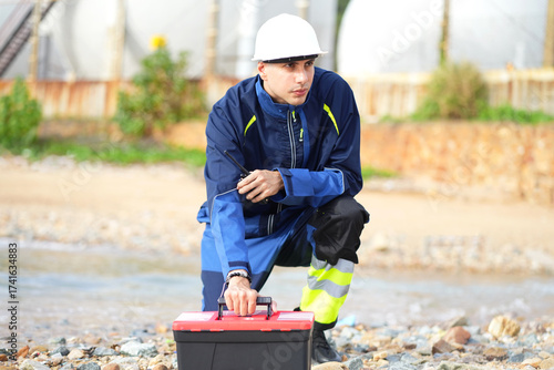 Environmental engineer or ecological scientist industrial worker in safety uniform working for inspect water quality at industry construction site near oil refinery plant by the sea.