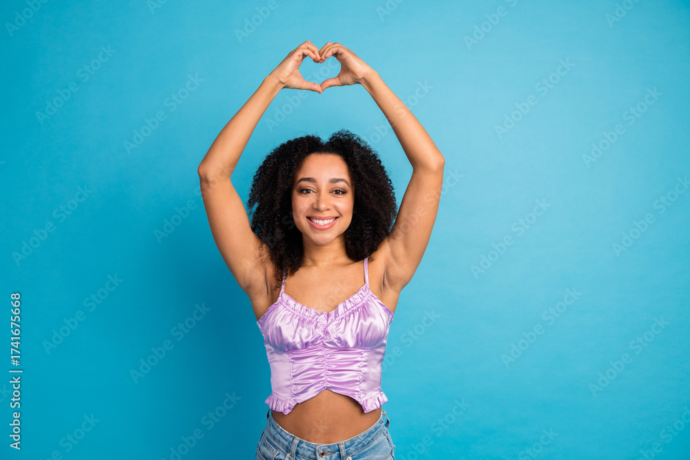 Fototapeta premium Young woman in a lavender top smiles and forms a heart with her arms against a bright blue background