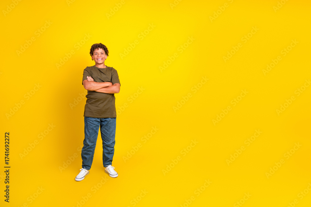 Fototapeta premium Funny confident young boy in casual khaki tshirt and jeans stands with arms crossed against a bright yellow background for a bold lifestyle promo