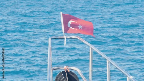 A Turkish flag (Ay Yıldız) proudly waving in the breeze on the railing of a boat or yacht. The background features the stunning, turquoise blue of the Mediterranean Sea. 