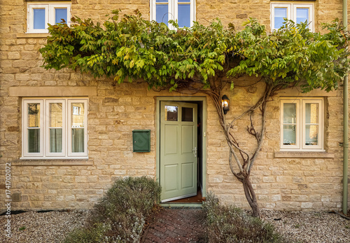 Traditional stone cottage entrance with open green door and leafy vine growing over wall. Cotswolds countryside, England
