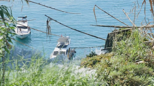 Waterfall plunging into the sea framed by green plants and bamboo. Two tour boats float in the turquoise water below. A unique coastal landscape of the Mediterranean. 