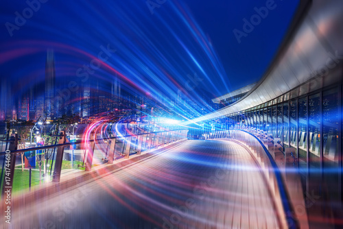 A creative nightscape of Hong Kong, China. Abstract streaks of light illuminate the elevated walkway against a dark sky, suggesting rapid transit and modern architecture. The creative composition show