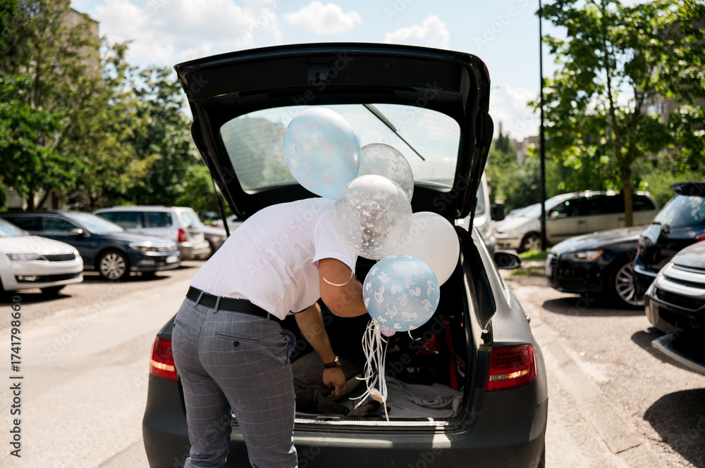 Fototapeta premium A person arranges blue and white balloons in the trunk of a car, preparing for a celebration on a sunny day in a parking lot surrounded by trees and parked vehicles.