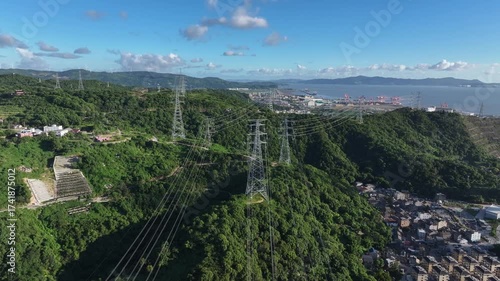 Transmission towers with power lines stand on a lush green hill under a blue sky