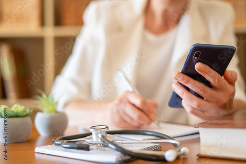 Healthcare professional performing medical consultation, making notes on paper, and holding a mobile phone, with a stethoscope on the desk, symbolizing modern primary care
