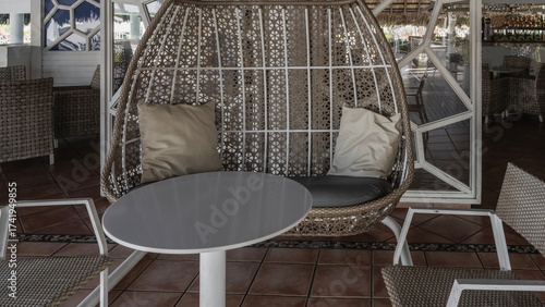 Wicker hanging swing chair on the cafe terrace. Cushions on the seat.  Round table, chairs nearby.