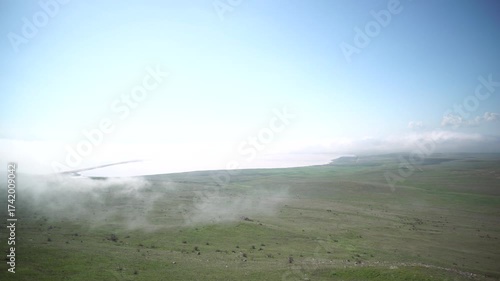 A foggy hillside with a clear blue sky in the background. The sky is dotted with clouds, and the grass is lush and green