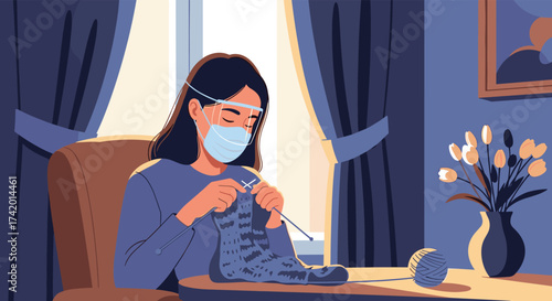A woman wearing a mask and face shield is knitting a sweater at home during the pandemic.
