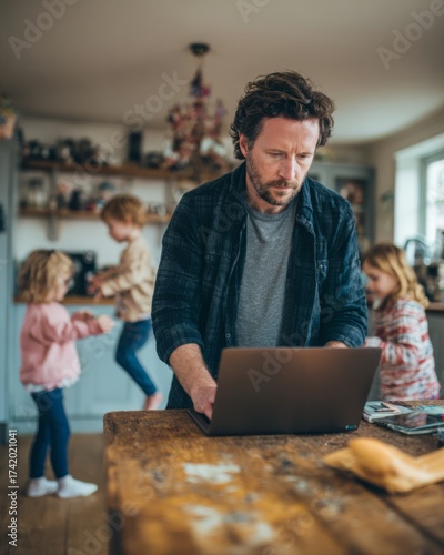 Family man working on a laptop on the kitchen table while children play around him.