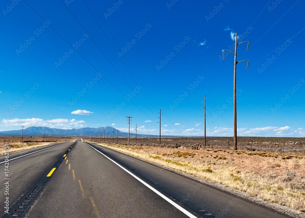 Fototapeta premium A straight desert highway in the American Southwest with uneven road markings, power lines, and distant mountains under a bright blue sky. Classic scenery of open road travel.