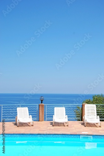 Minimalist summer scene with white deck chairs by a swimming pool overlooking the calm blue sea.