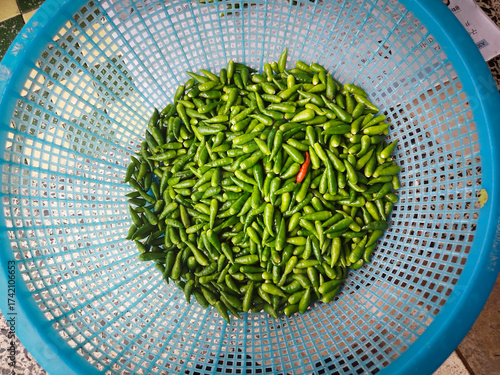 A single orange-red chili pepper stands out prominently amidst a dense, full-frame pile of vibrant green chili peppers.