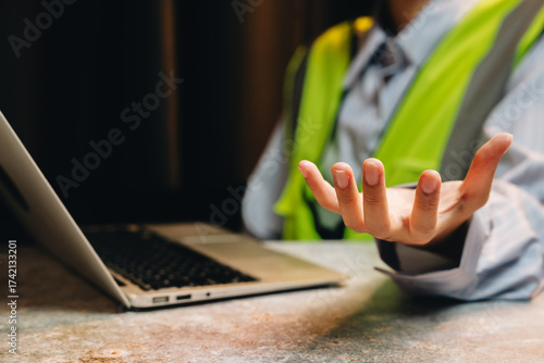 Professional construction worker in reflective vest gestures while consulting a laptop during a project meeting indoors