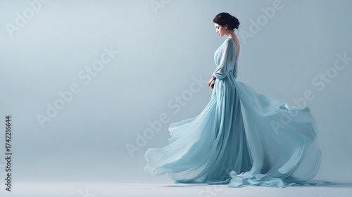 A woman in an airy, flowing blue gown stands with a subtle pose, looking down, against a pastel backdrop