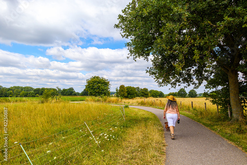 Rural road with adult woman walking with dog among farmland, wearing casual clothes and hat, back to camera, green leafy trees in background, cloudy summer day in Limburg, Netherlands