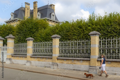 Senior woman walking with dachshund on sidewalk, brick wall with wrought iron outside the castle, leafy green trees, sloping hipped roof with chimneys in background in Rekem, Limburg, Belgium