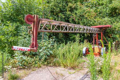 Old, obsolete tractor with an agricultural crane, abandoned among lush wild vegetation of farm in agricultural land, rusty red metal arm, leafy trees in background, sunny spring day