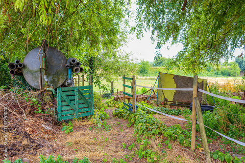 Corral field plot in agricultural farm, old water storage tank with pipes and hoses, green wooden gate, fences with wooden posts, planks, abundant leafy trees in background, sunny spring day