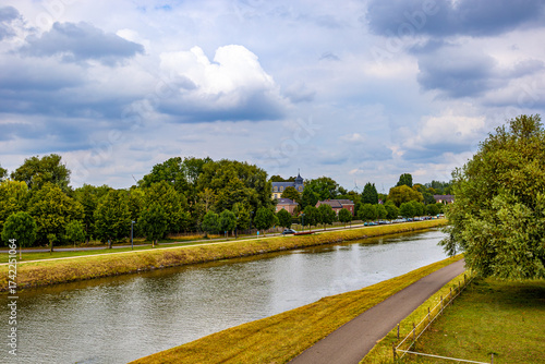 Canal landscape between two country roads, green leafy trees and houses in background against stormy sky, cars parked on canal bank, cloudy day on outskirts of Oud-Rekem, Limburg, Belgium