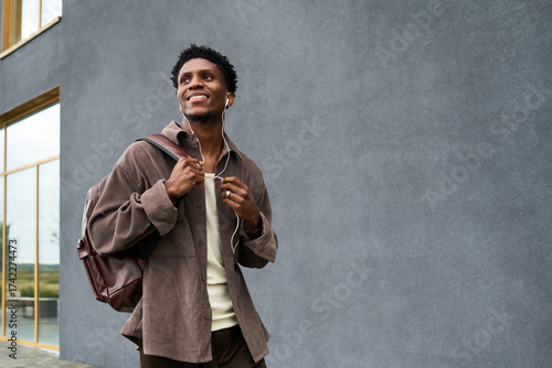 Fotografie Young Black man walking outdoors carrying backpack and listening to music with e