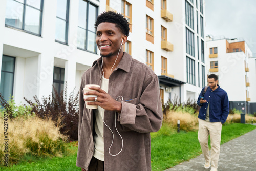Young Black man walking outdoors holding coffee cup and listening to music with earphones, smiling while passing by young man using smartphone in modern urban area