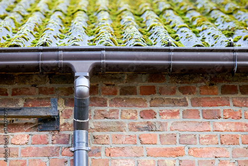 A brown plastic gutter and downspout on a rustic brick wall, below a mossy, corrugated roof. Highlights exterior building maintenance needs.