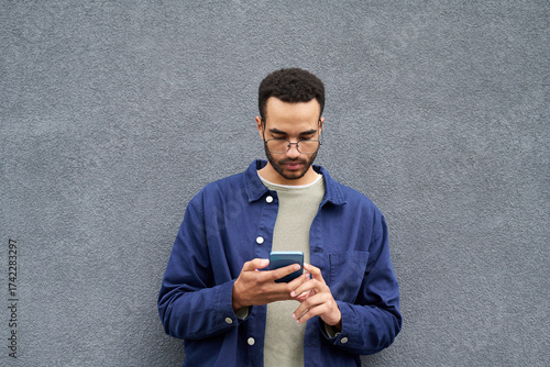 Photos Young Black man standing against plain gray wall using smartphone, wearing glass
