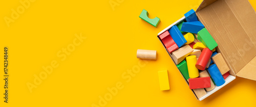 Wooden children's multi-colored bright construction set blocks in cardboard box on yellow background. Montessori style cubes game for toddlers flat lay top view. Concept of learning and education