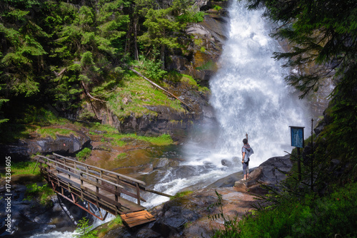 Traveler Stands on Wet Rocks Beside a Tall Waterfall and Wooden Bridge in the Forest, 
A lone traveler stands on a rocky ledge beside a powerful waterfall, pointing toward the spray. 