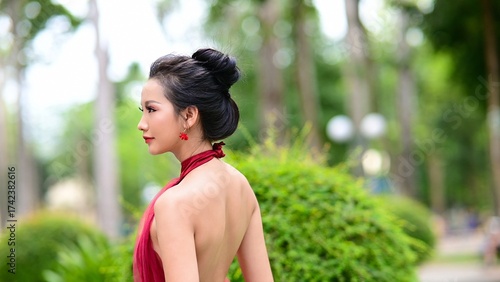 Elegant Asian woman in red backless dress posing gracefully in green park under natural sunlight