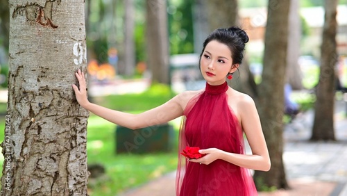 Elegant Asian woman in red backless dress posing gracefully in green park under natural sunlight