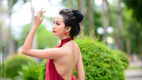 Elegant Asian woman in red backless dress posing gracefully in green park under natural sunlight