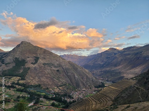 A imagem retrata o Vale Sagrado dos Incas ao entardecer, com montanhas imponentes banhadas pela luz dourada do sol e nuvens alaranjadas no horizonte. No sopé das montanhas, observa-se um pequeno vilar
