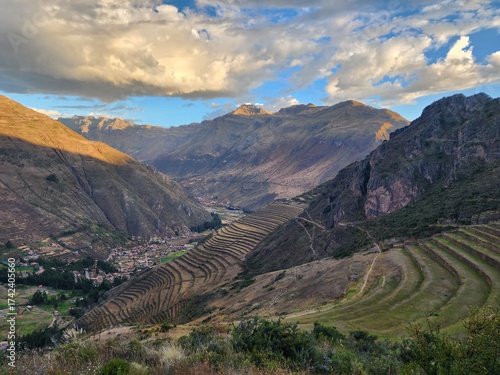 A imagem retrata o Vale Sagrado dos Incas ao entardecer, com montanhas imponentes banhadas pela luz dourada do sol e nuvens alaranjadas no horizonte. No sopé das montanhas, observa-se um pequeno vilar