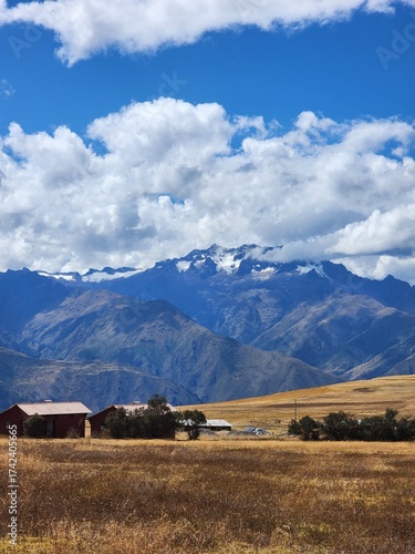 A imagem mostra uma paisagem rural andina com campos dourados e algumas casas simples em primeiro plano. Ao fundo, erguem-se montanhas majestosas da Cordilheira dos Andes, parcialmente cobertas por ne