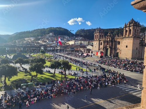 A imagem mostra a Plaza de Armas de Cusco, repleta de pessoas reunidas para um evento festivo sob um céu azul intenso. Ao fundo, destaca-se a imponente Catedral de Cusco, construída em pedra no estilo
