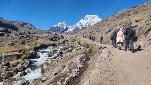 A imagem mostra um grupo de pessoas caminhando por uma trilha nas montanhas andinas sob um céu azul límpido. Ao lado do caminho, um riacho de águas cristalinas desce entre pedras e vegetação rasteira.