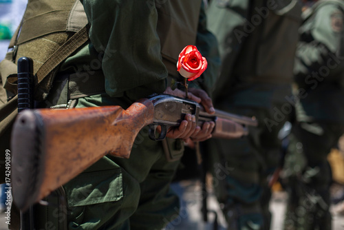A Venezuelan soldier from behind holds a rose, a symbol of violence and peace, next to his weapon.