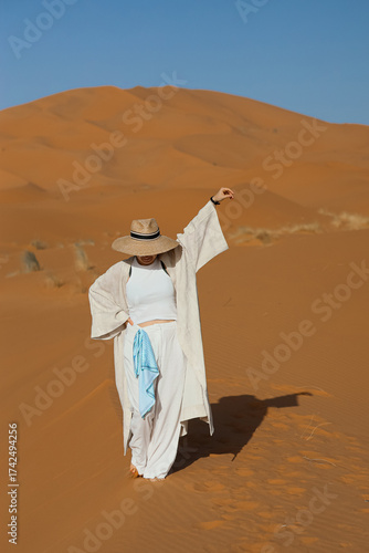 A woman walks through the desert wearing a hat and a white dress