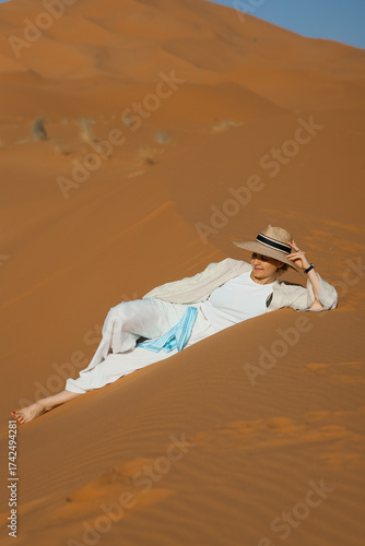 A woman in white clothes and a hat is sitting on a desert dune