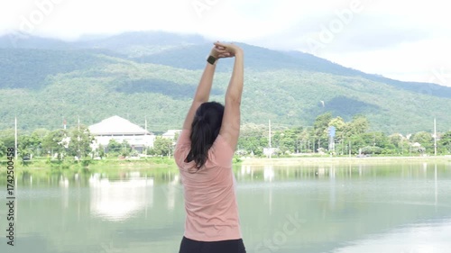 Asian woman stretches her arms above her head, performing a pre-run warm-up in a scenic suburb. The background features stunning mountain views and a peaceful reservoir.