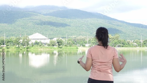 Asian woman shakes her hands and stretches her arms above her head, performing a pre-run warm-up in a scenic suburb. The background features stunning mountain views and a peaceful reservoir.
