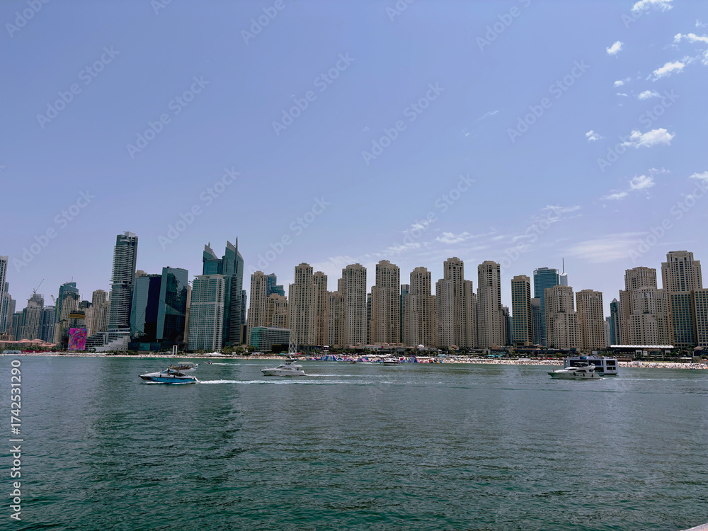 Obraz premium Modern city skyline with tall skyscrapers by the sea and boats on the water under a clear blue sky, urban waterfront cityscape on a sunny day