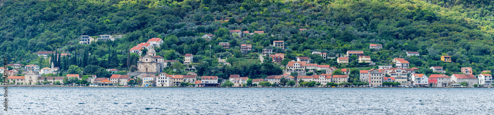 Fototapeta premium Panoramic view of Prcanj, Kotor, Montenegro, along Jadranska magistrala with historic stone houses, church domes, and green hills rising above the calm Adriatic Sea.