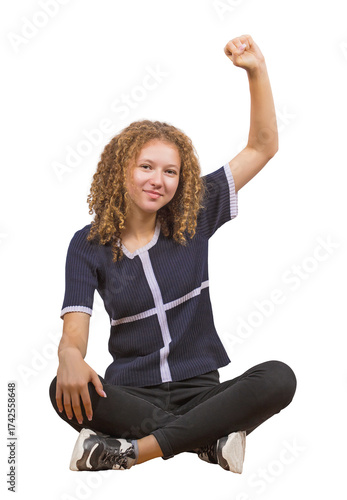 Full length portrait of curly teenage girl sitting carefree on the floor, raising up her fist looking positive to camera isolated on transparent background. Seated frizzy young woman, adolescent