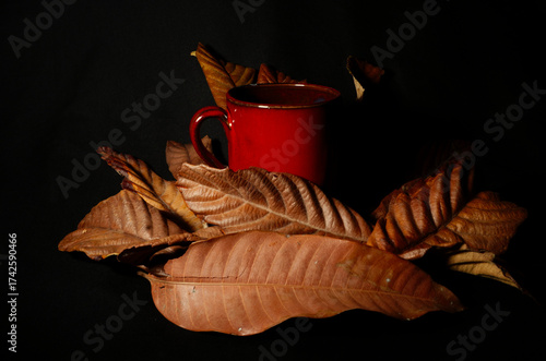 Autumn still life with red cup among dry leaves on dark background. Warm seasonal mood evoking fall evenings and cozy atmosphere, also suitable for Halloween themes.