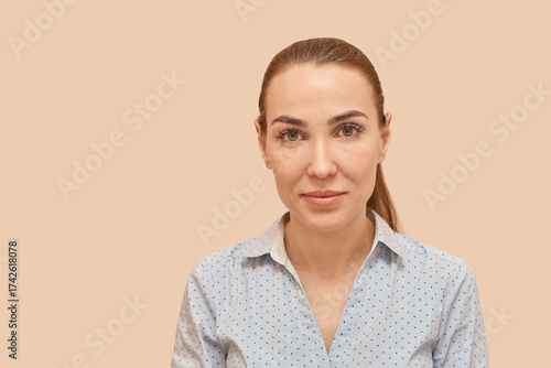 Young caucasian female in polka dot shirt with neutral expression on beige background.