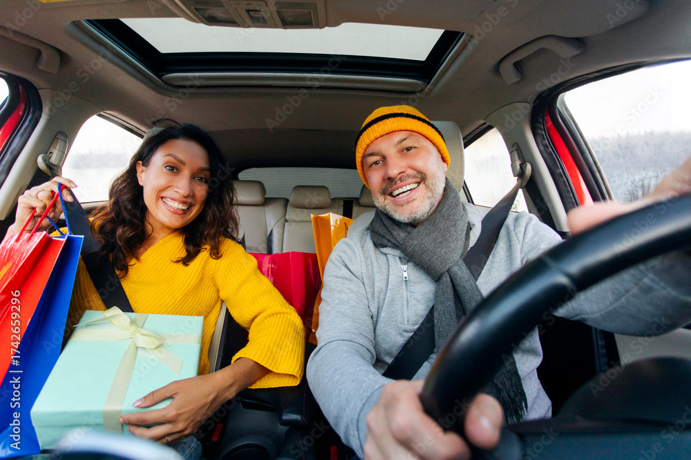 Obraz premium Happy couple driving car full of presents after buying gifts in mall, smiling at camera. Christmas preparation, seasonal sale